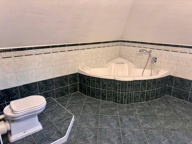 Badezimmer im Dachgeschoss mit Eckbadewanne und dunklen Fliesen, unter einer Dachschräge.