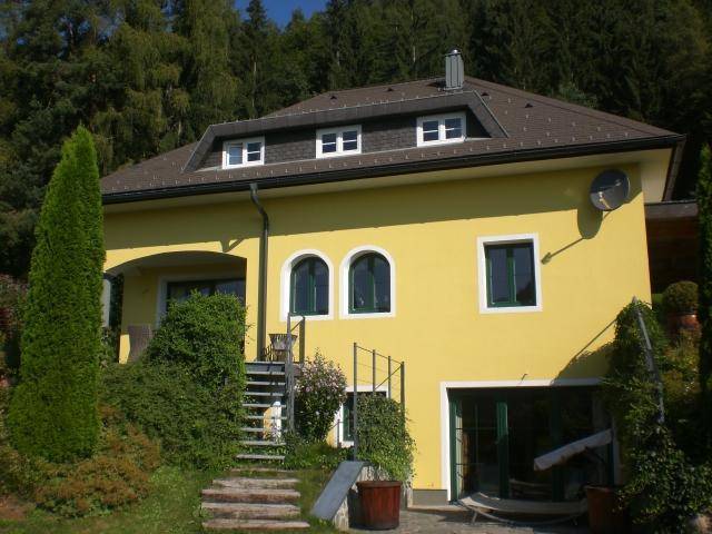 Gelbes Haus mit Dachgauben, Außentreppe und Garagenzugang, umgeben von Grün.