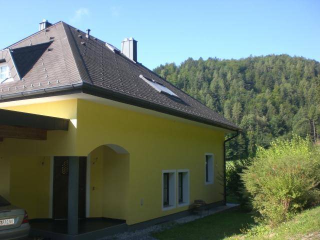 Gelbes Haus mit Carport und Satteldach, umgeben von Bäumen und Sträuchern.