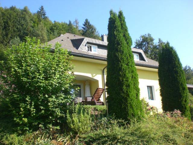 Gelbes Haus mit kleiner Terrasse und Gartenmöbeln, umgeben von üppiger Vegetation.