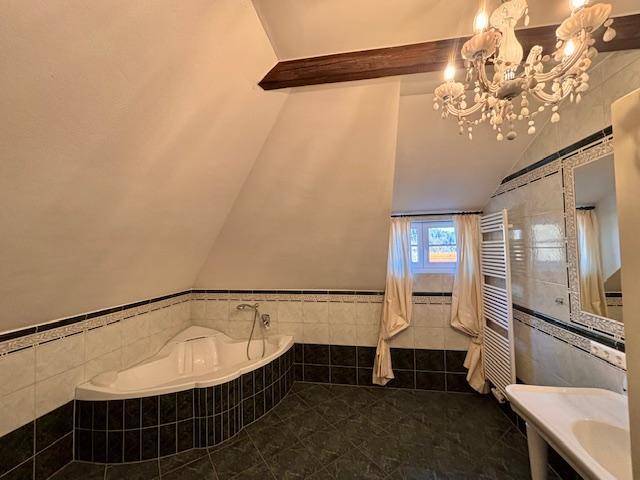 Helles Badezimmer mit Eckbadewanne, Fenster und beheiztem Handtuchhalter unter Dachschräge.