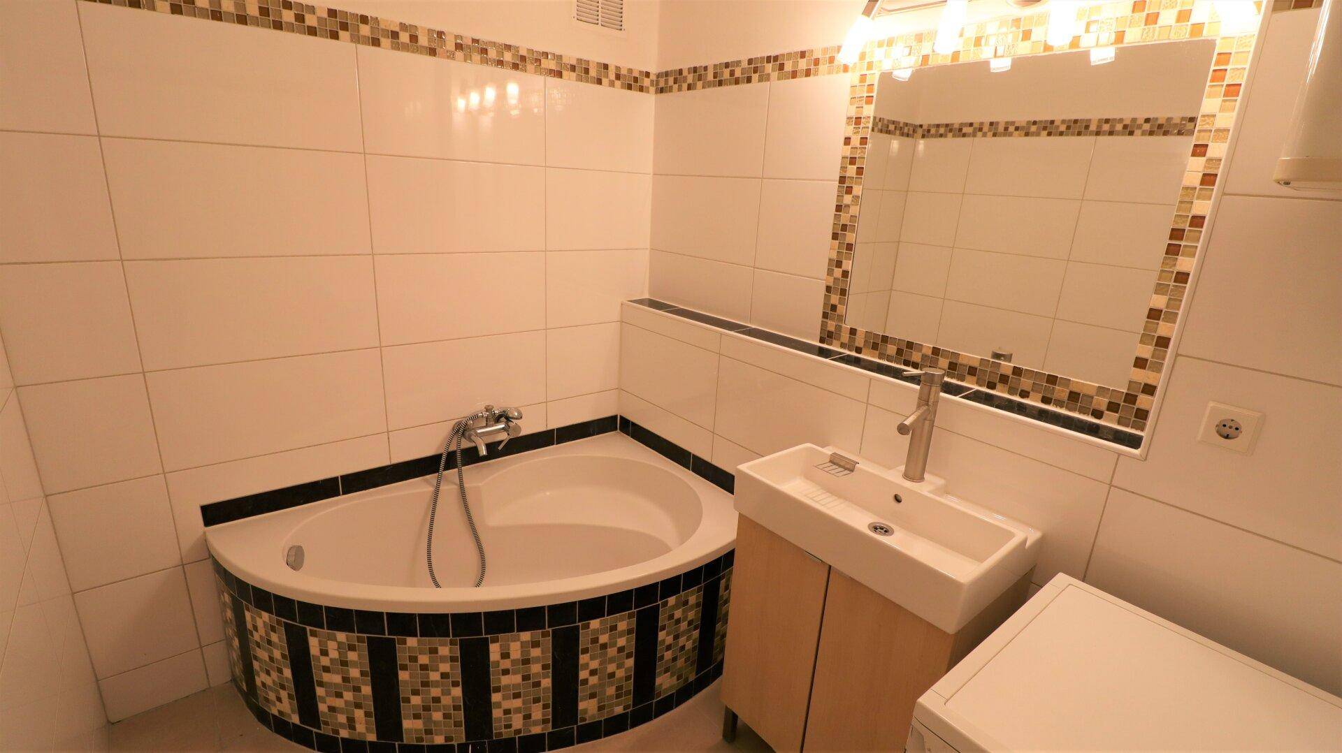Modern gefliestes Badezimmer mit Eckbadewanne, Waschbecken und Spiegel mit Beleuchtung.