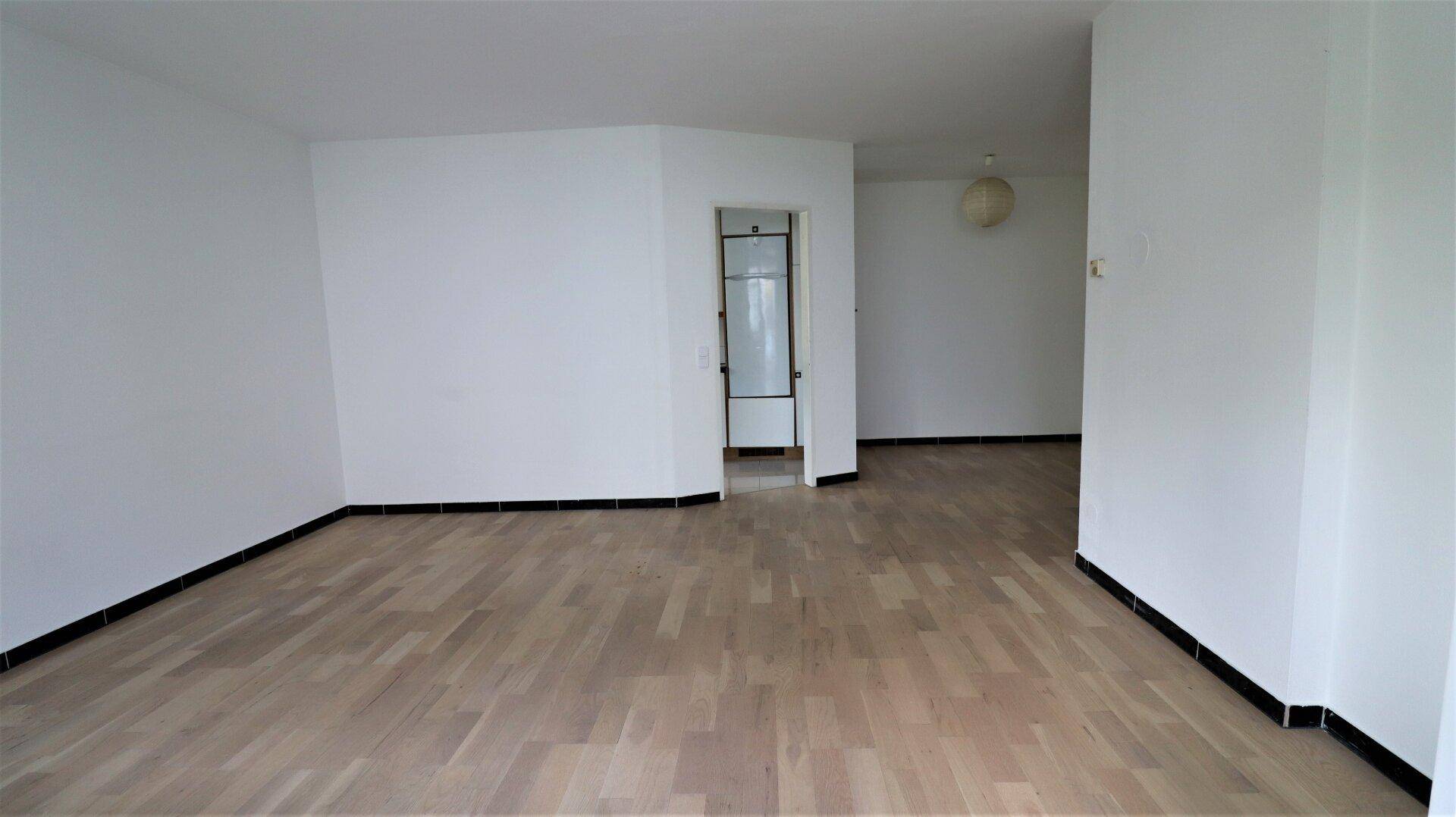 Geräumiger, leerer Wohnbereich mit hellem Holzfußboden und Zugang zu weiteren Räumen.