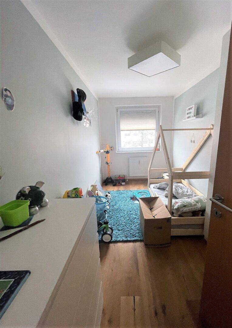 Kinderzimmer mit Holzbettgestell, blauem Teppich, weißer Kommode und Spielzeug, hell durch ein Fenster.