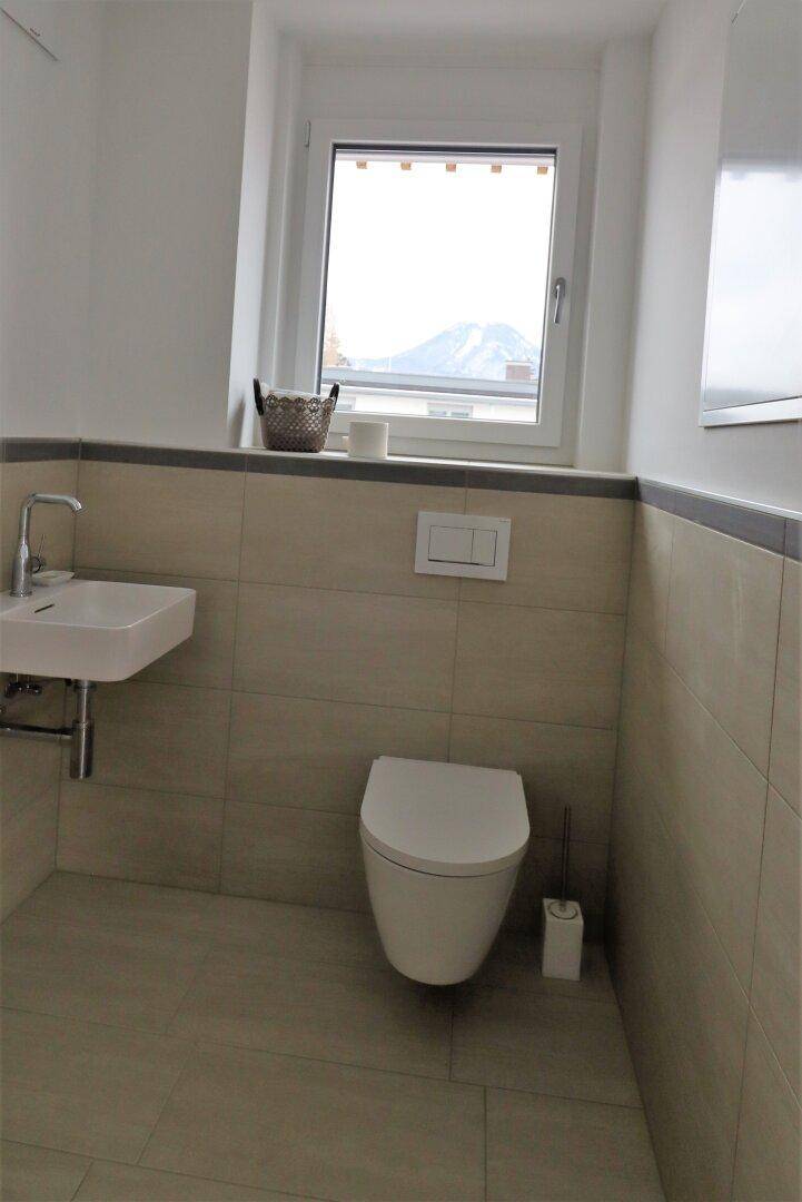 Helles Gäste-WC mit kleinem Waschbecken, wandhängendem WC und Fenster mit Bergblick.