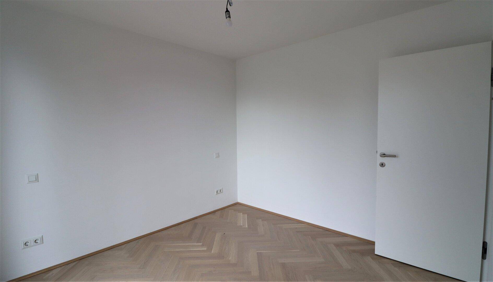 Leeres Zimmer mit hellem Fischgrätparkett und weiß gestrichenen Wänden.