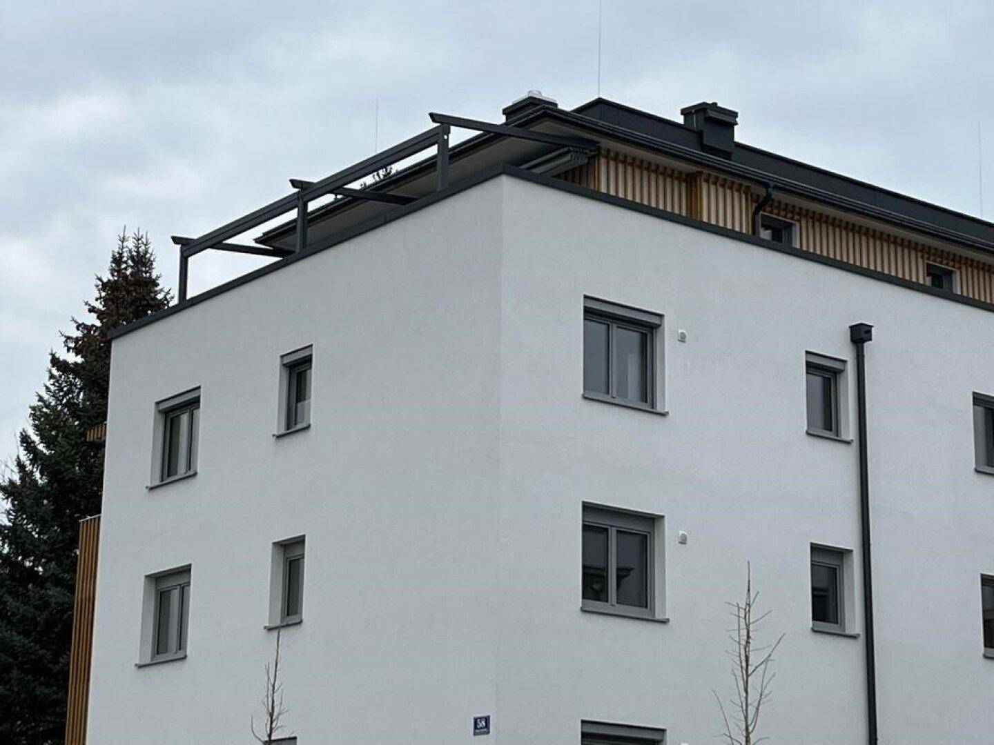 Moderne Gebäudefassade mit weißen Wänden, grauen Fenstern und Holzelementen im Dachbereich.