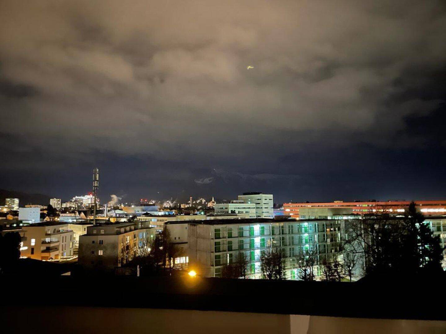 Nächtlicher Blick auf die beleuchtete Stadt mit Bergen im Hintergrund unter bewölktem Himmel.