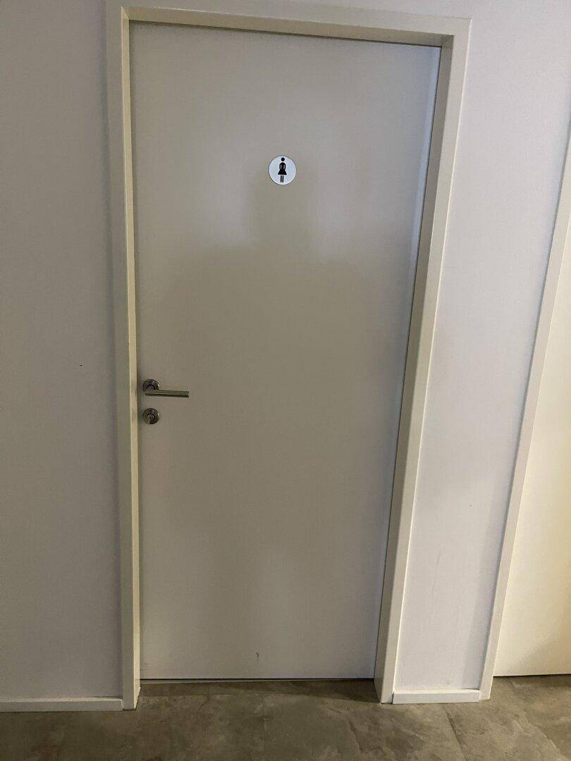 Eine schlichte weiße Tür mit einem Damensymbol, die zu einer Toilette führt.