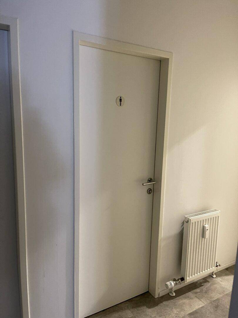 Eine schlichte weiße Tür mit einem Herrensymbol, die zu einer Toilette führt, daneben ein Heizkörper.
