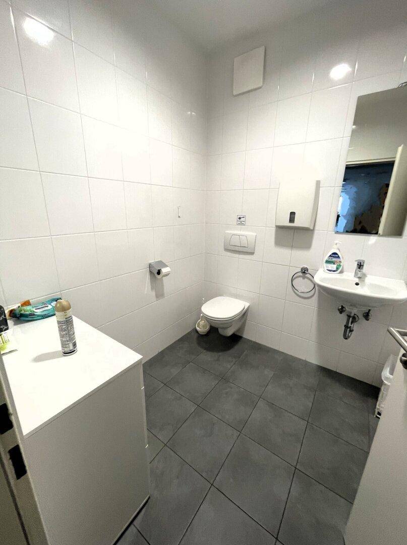 Saubere, geflieste Toilette mit Waschbecken, Spiegel und Wand-WC, funktional und hell.