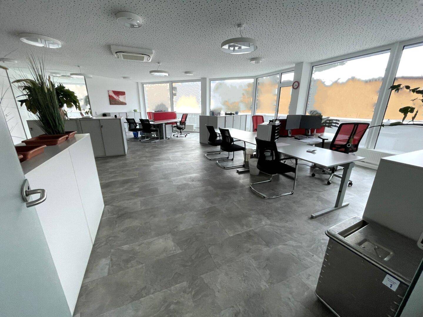 Heller Großraumbüro mit mehreren Arbeitsplätzen, großen Fenstern und modernen Büromöbeln.