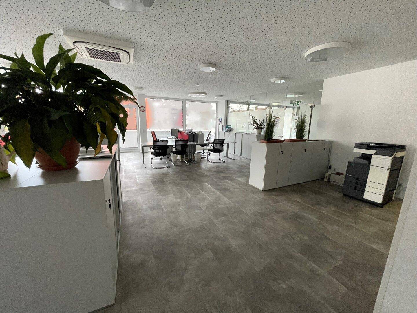 Moderner Bürobereich mit Schränken, Arbeitsplätzen und einem Kopierer, gut beleuchtet durch große Fenster.