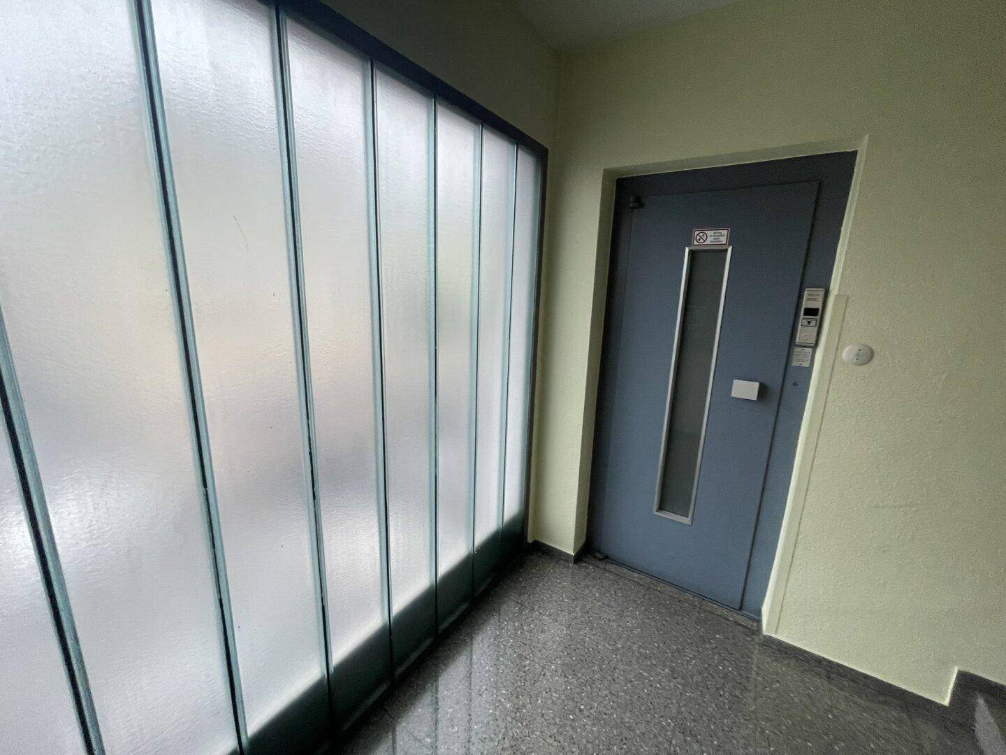 Heller Flur mit Milchglaswand und einer grauen Eingangstür, die zu einem Aufzug führt.