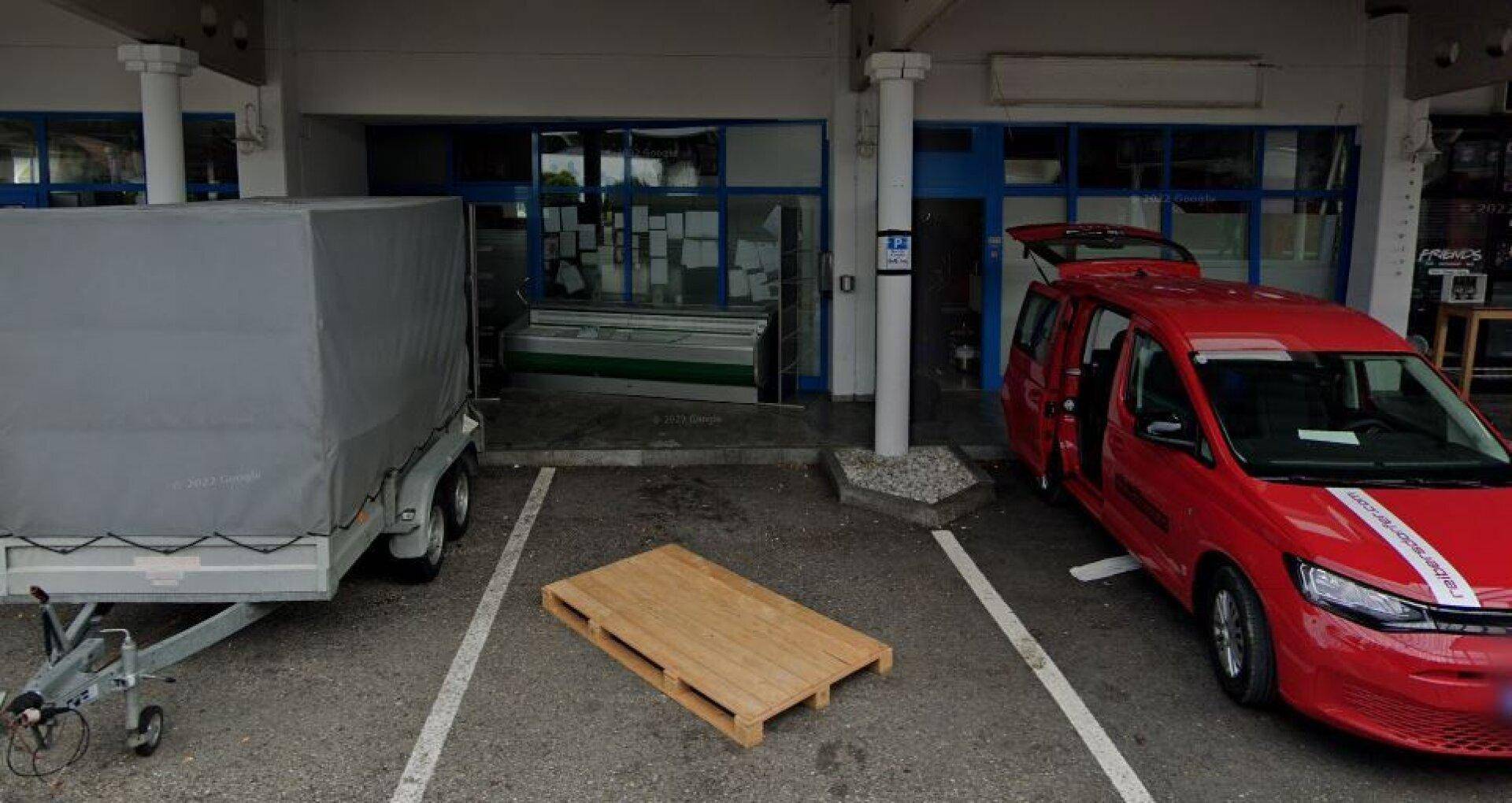 Geschäftsfassade mit blauen Fensterfronten und einem Parkplatz, auf dem ein Anhänger und ein roter Lieferwagen stehen.