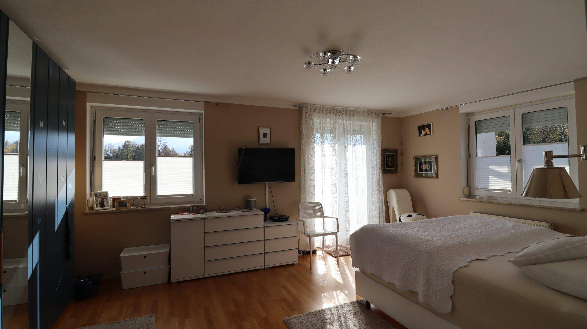 Schlafzimmer mit Fernseher, Kommode und zwei Fenstern für viel Tageslicht.