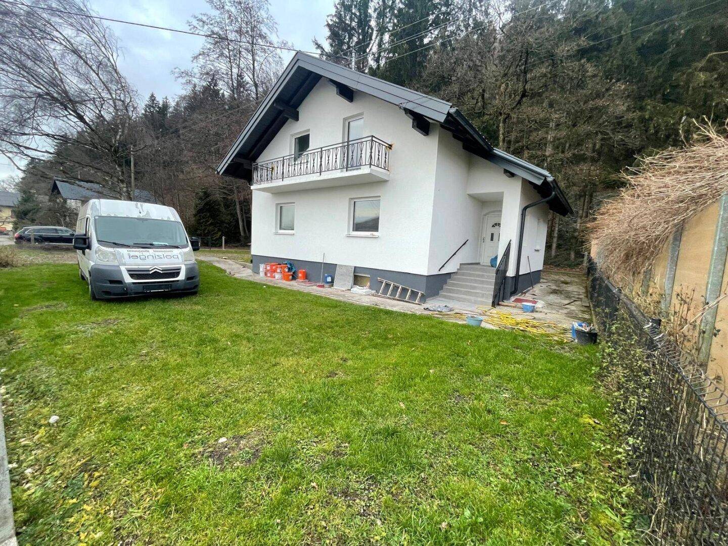 Freistehendes Haus mit weißer Fassade und Balkon, umgeben von einem großen Rasen und Wald im Hintergrund.