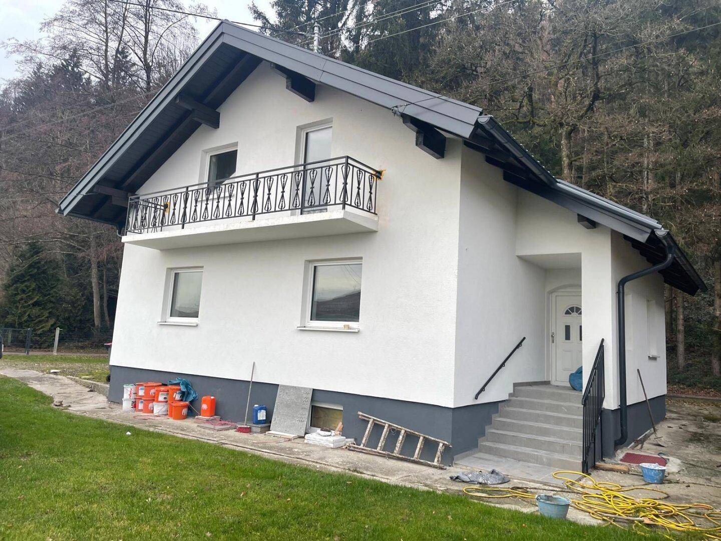 Nahaufnahme des frisch gestrichenen weißen Hauses mit dunklem Dach, Balkon und Eingangsbereich, umgeben von Natur.