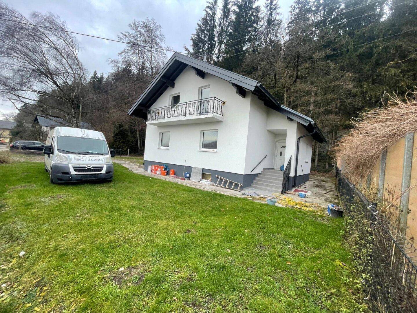 Modernes Einfamilienhaus mit weißer Fassade, dunklem Dach und kleinem Balkon, umgeben von Grünflächen.