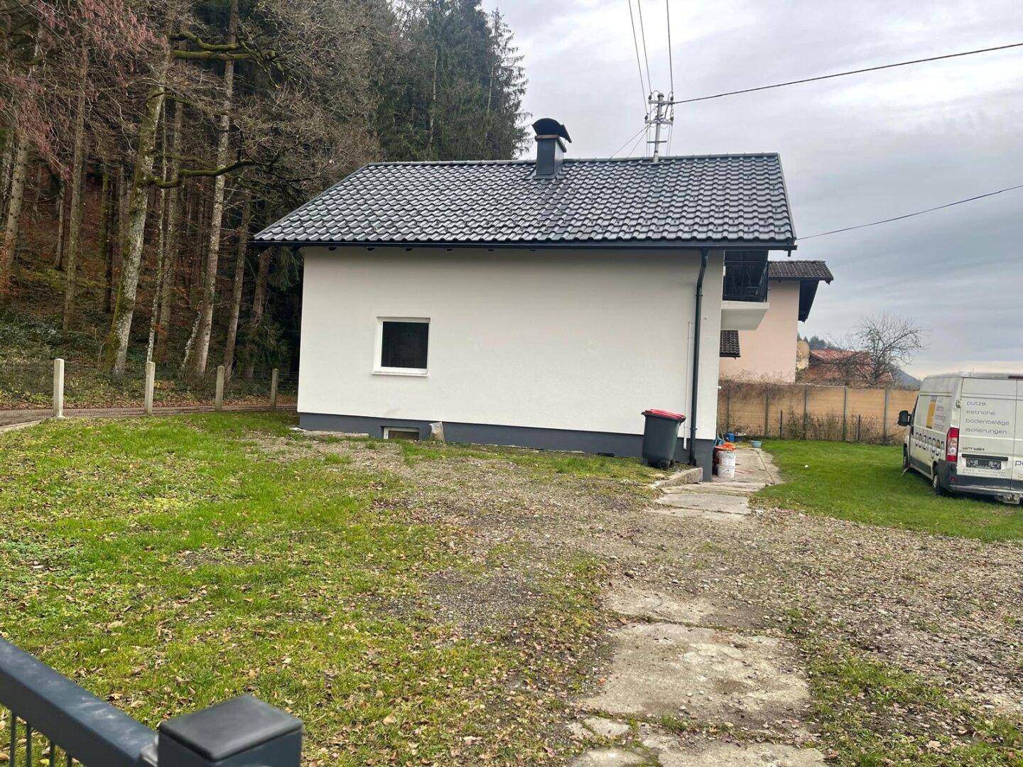 Seitenansicht des Hauses mit Garten und Zufahrt, umgeben von Bäumen und ländlicher Umgebung unter bewölktem Himmel.