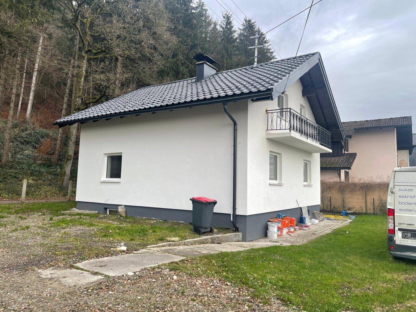 Seitenansicht des Hauses mit Garten und Zufahrt, umgeben von Bäumen und ländlicher Umgebung unter bewölktem Himmel.