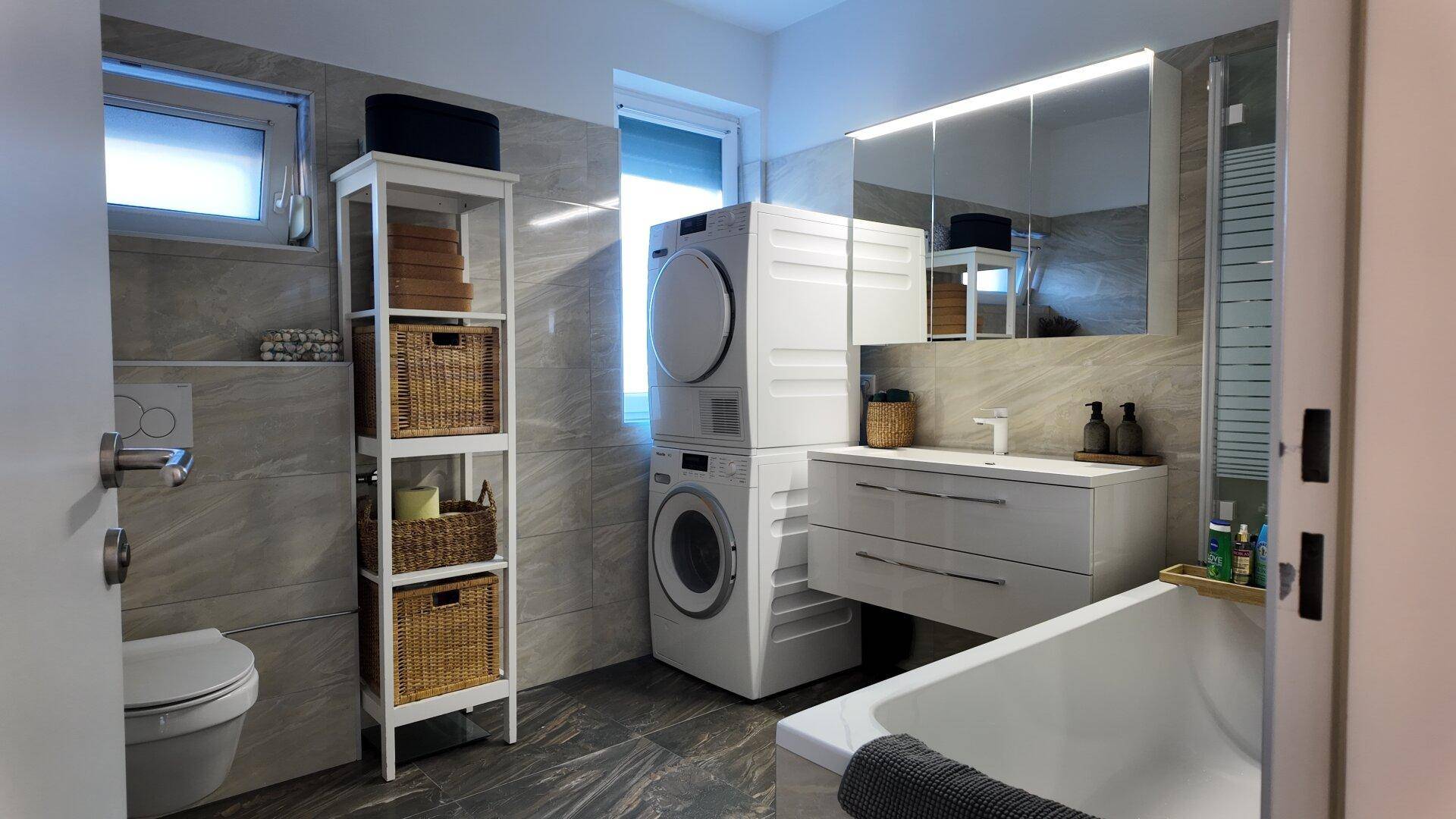 Modernes Badezimmer mit Badewanne, Waschbecken, WC und integriertem Waschmaschinen-Trockner-Turm.