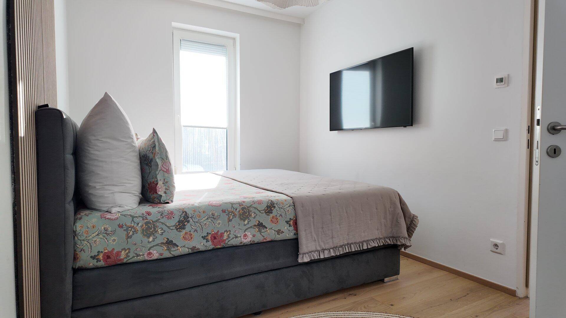 Modern eingerichtetes Schlafzimmer mit Wandfernseher und viel Tageslicht durch das große Fenster.