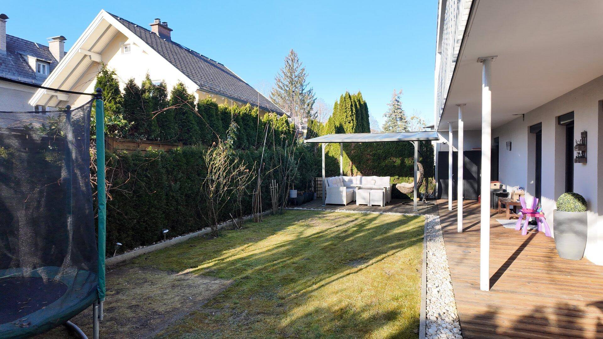Gepflegter Garten mit Rasenfläche, Trampolin und überdachtem Sitzbereich auf einer Holzterrasse.