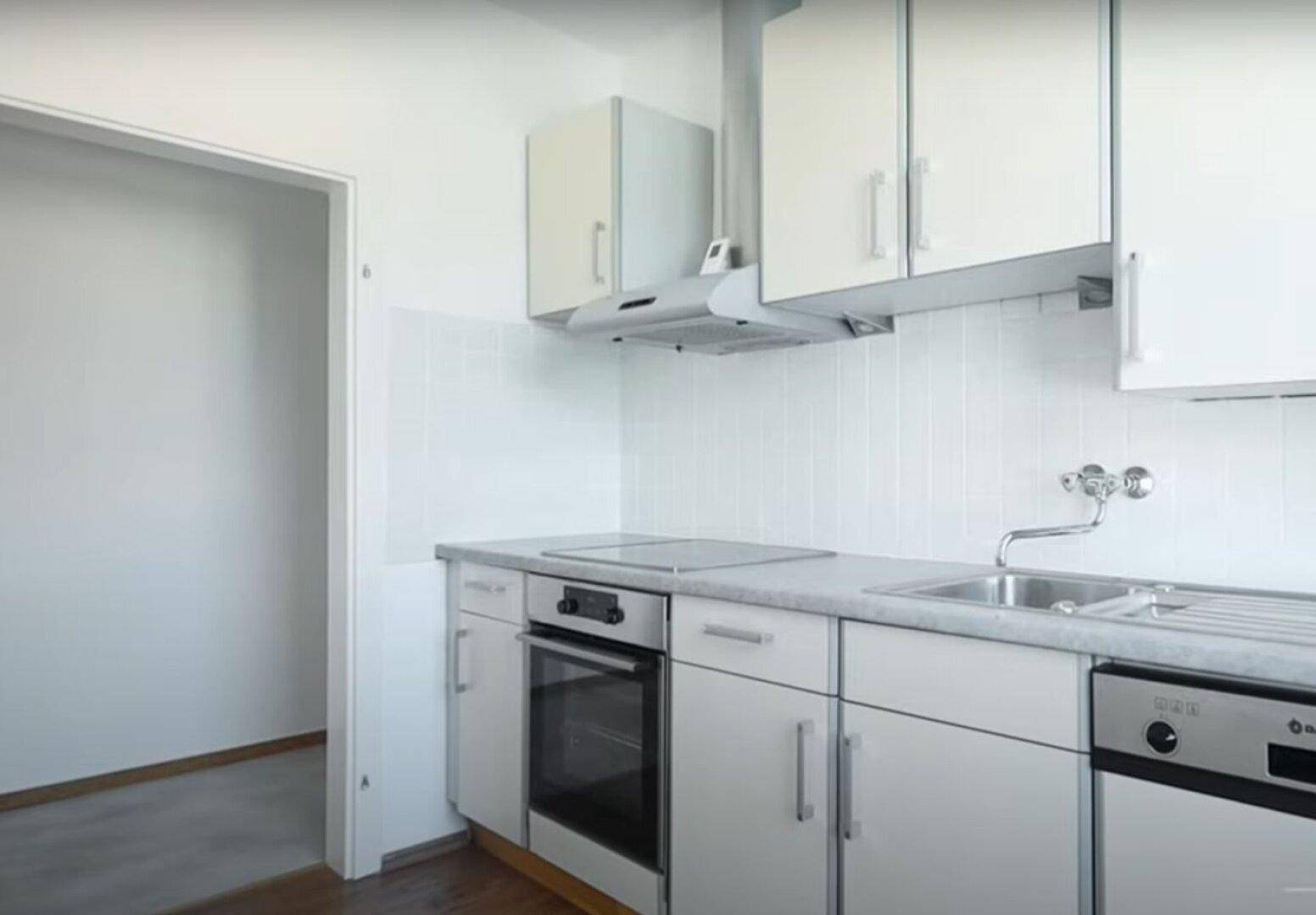 Helle Einbauküche mit weißen Schränken, Spüle, Kochfeld und Backofen, angrenzend an einen leeren Raum.
