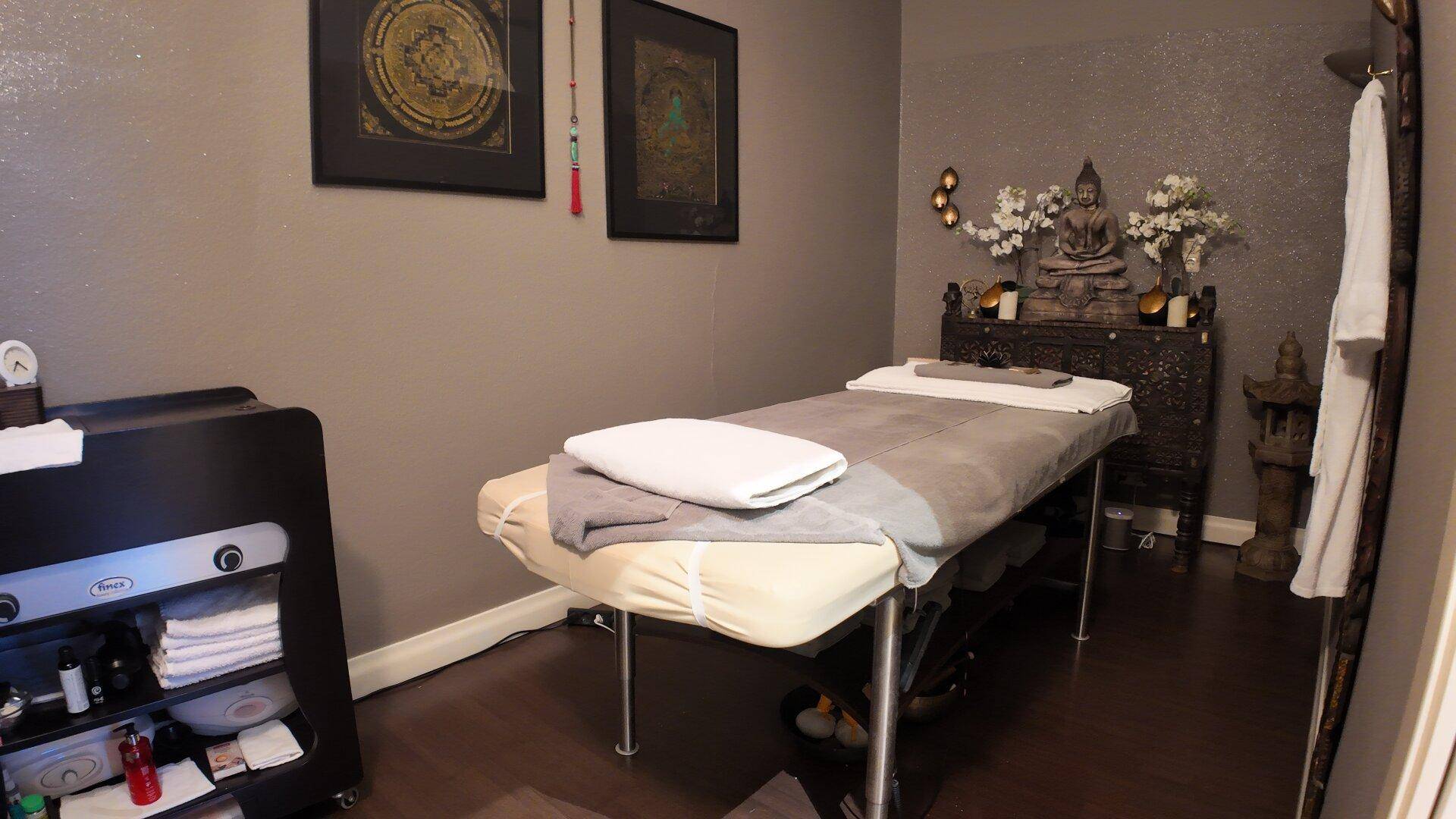 Ruhiger Massageraum mit einer Liege, asiatischer Dekoration und dunklem Holzboden.