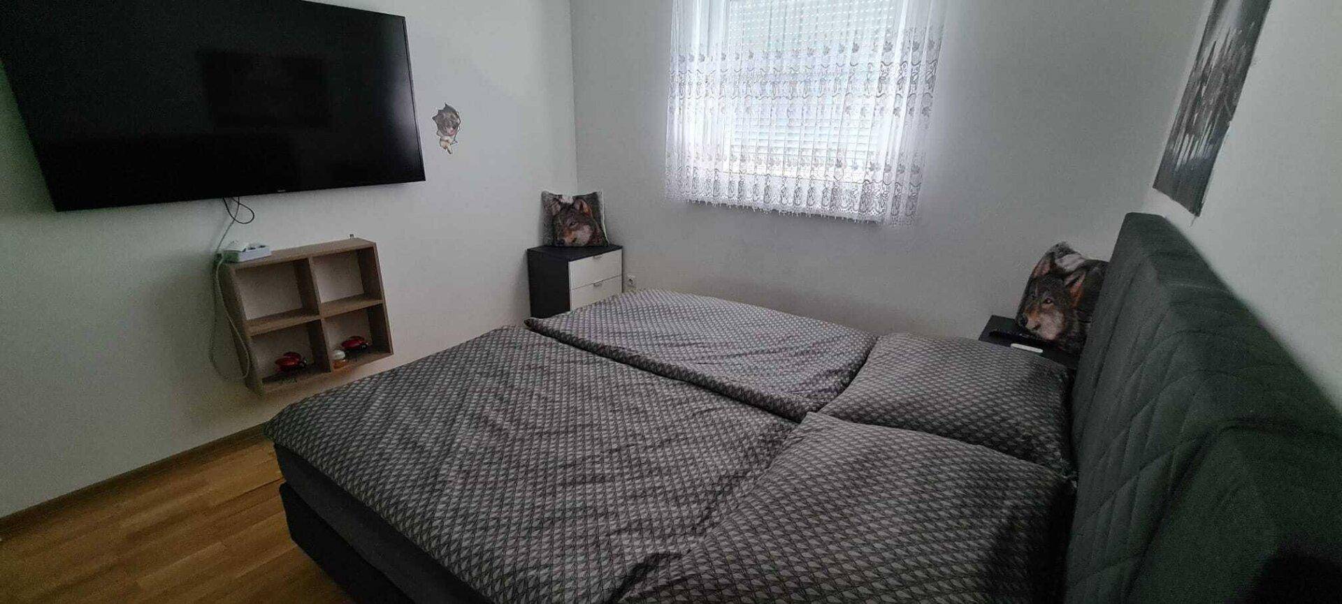 Gemütliches Schlafzimmer mit Doppelbett, Wandfernseher und Fenster mit Vorhängen.