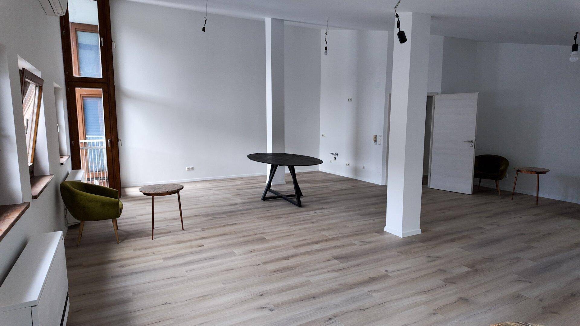 Heller, offener Raum mit modernem Laminatboden, weißen Wänden und großen Fenstern.