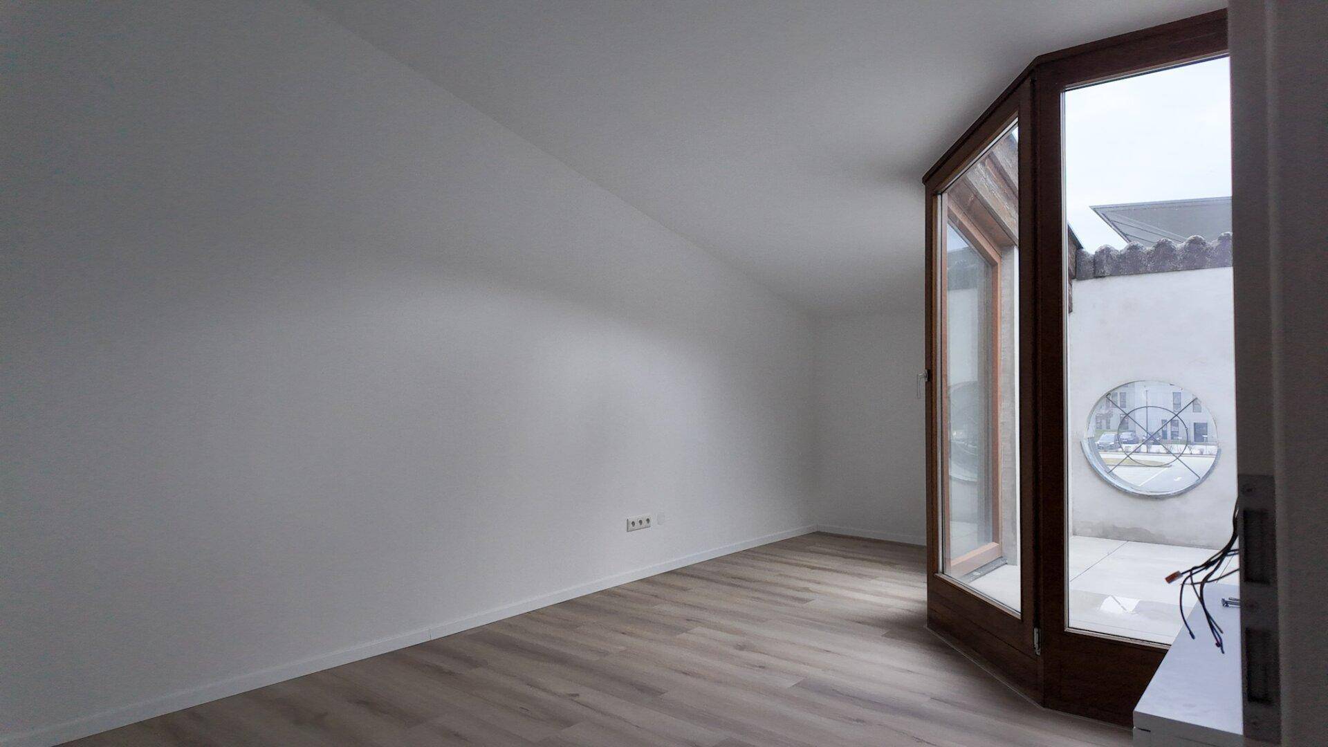Heller Raum mit Laminatboden und einer großen Holztür, die zu einer Außenterrasse führt.