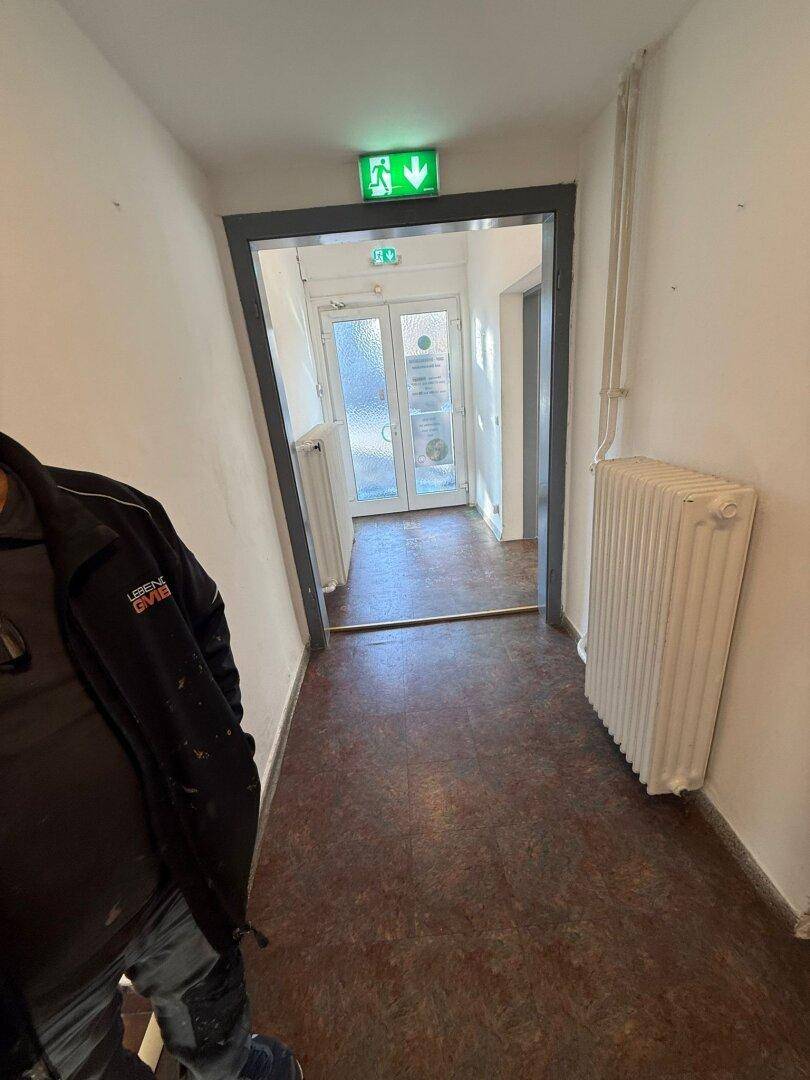 Flur führt zu einem Ausgang mit doppelten Glastüren und einem grünen Notausgangsschild.