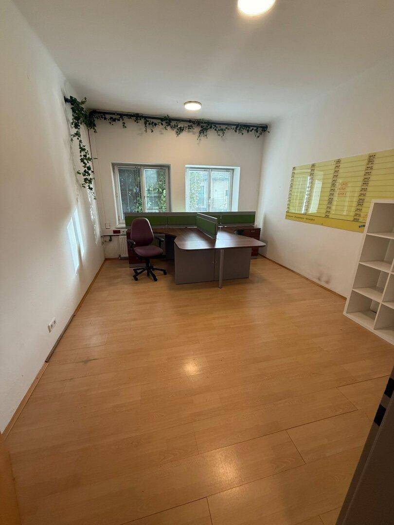 Büroraum mit einem Schreibtisch, Bürostuhl, Regalen und Fenstern für natürliches Licht.