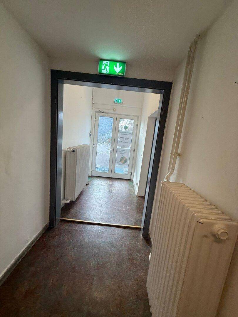 Flur führt zu einem Ausgang mit doppelten Glastüren und einem grünen Notausgangsschild.
