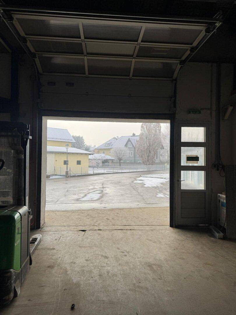 Blick aus der Lagerhalle durch das offene Rolltor auf eine verschneite Straße und Wohnhäuser. Die Umgebung ist ländlich geprägt.
