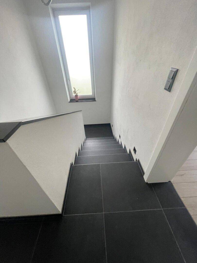 Treppenabgang mit dunklen Fliesenstufen und einem hohen, schmalen Fenster für Helligkeit.