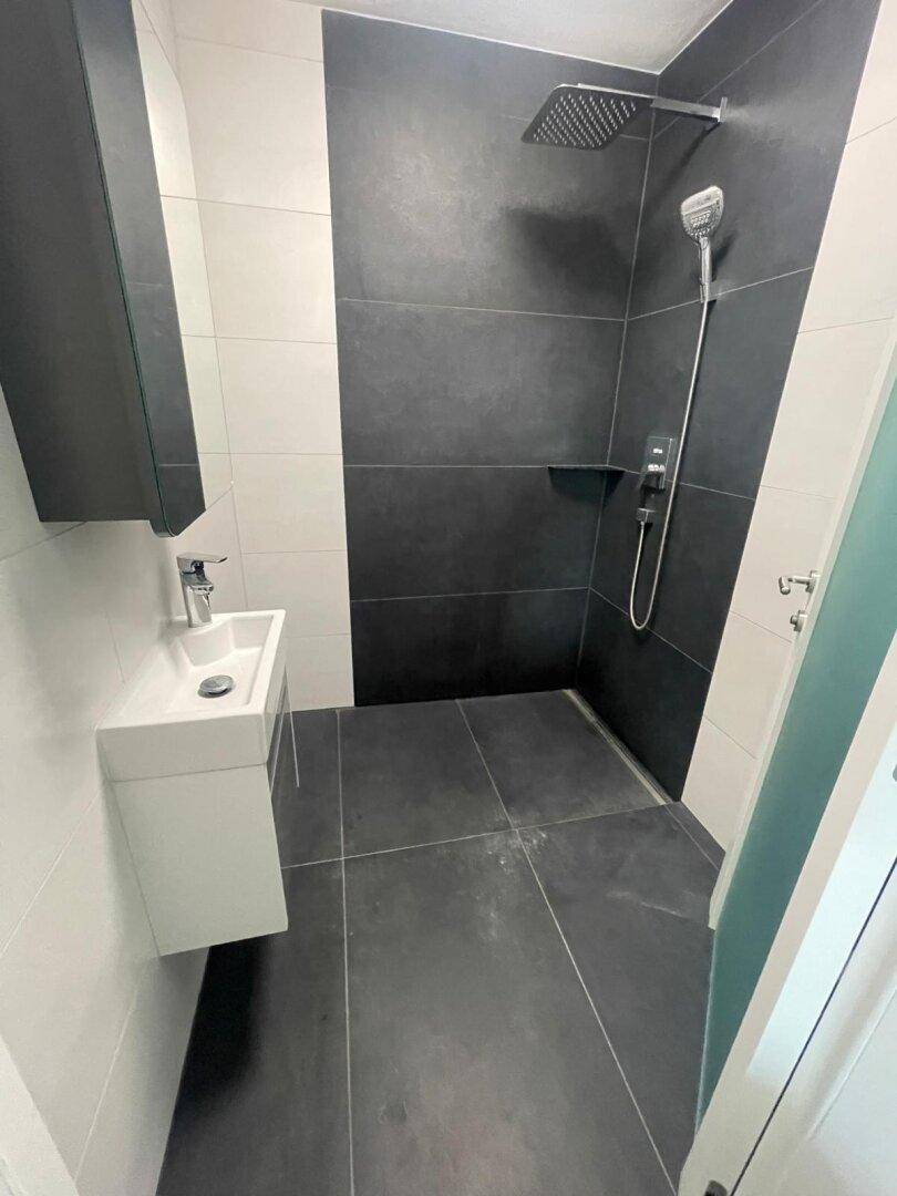 Modernes Gäste-WC mit Dusche, kleinem Waschbecken und dunklen Fliesen an Wänden und Boden.