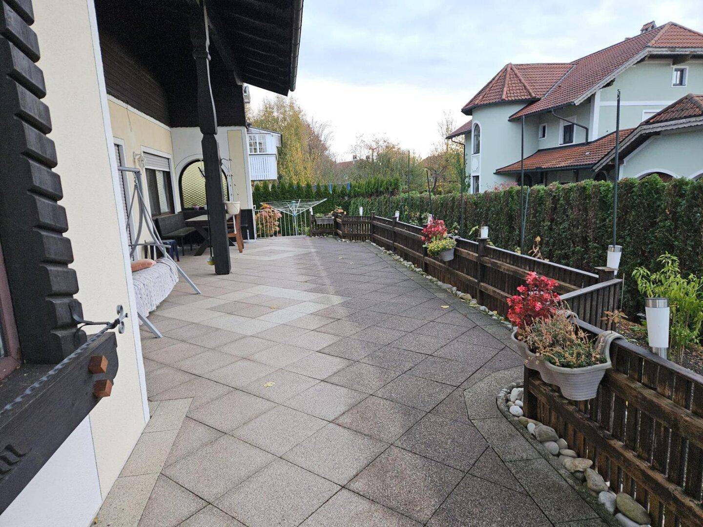 Lange, gepflasterte Terrasse entlang des Hauses mit Blumenkästen und Holzzaun.