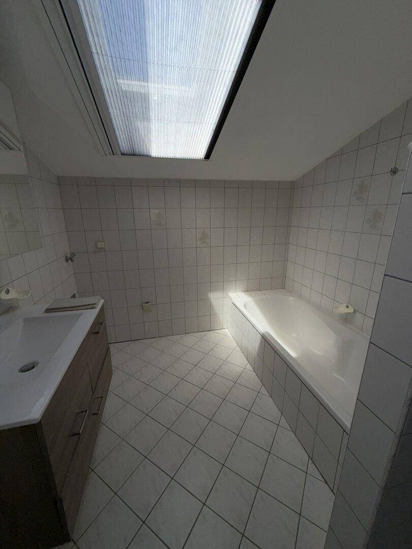 Helles Badezimmer mit Badewanne, Waschtisch und großem Dachfenster, das viel Tageslicht hereinlässt.