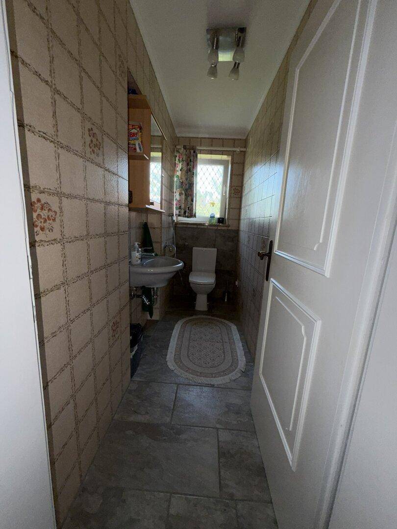Badezimmer mit beigefarbenen Fliesen, Waschbecken, Toilette und Fenster für Tageslicht.