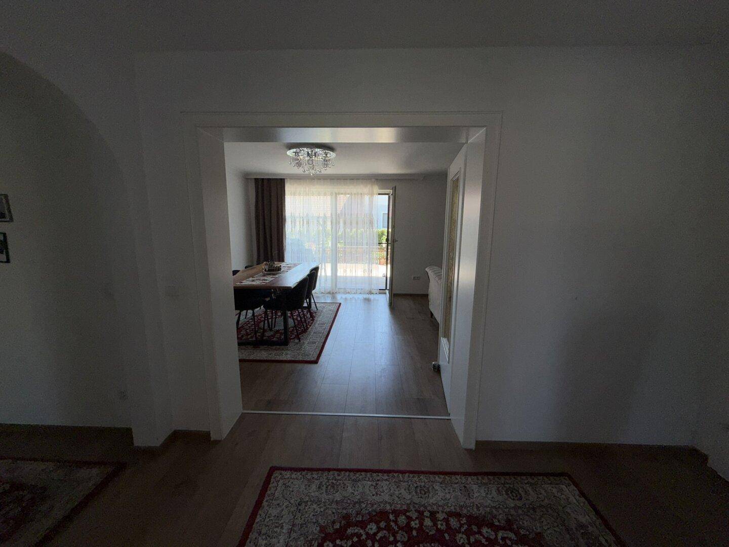 Blick durch einen offenen Durchgang in den hellen Essbereich mit Holzboden und Zugang zum Außenbereich.
