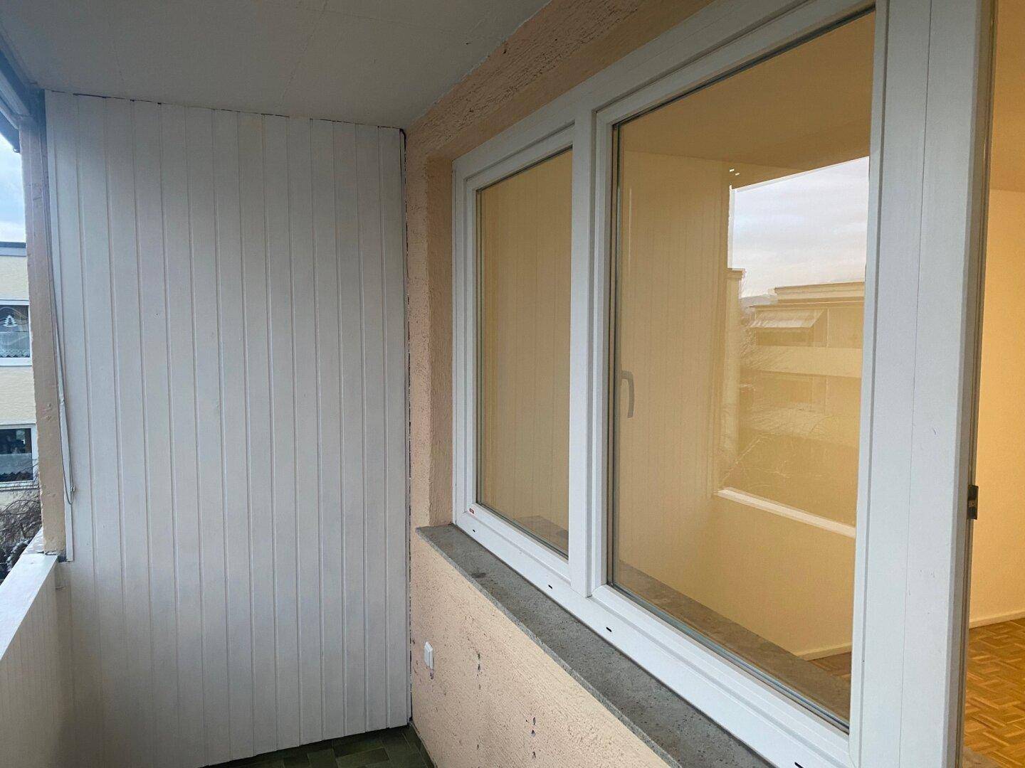 Balkon mit weißer Verkleidung und Fenster, das den Innenraum widerspiegelt, mit Blick auf die Umgebung.