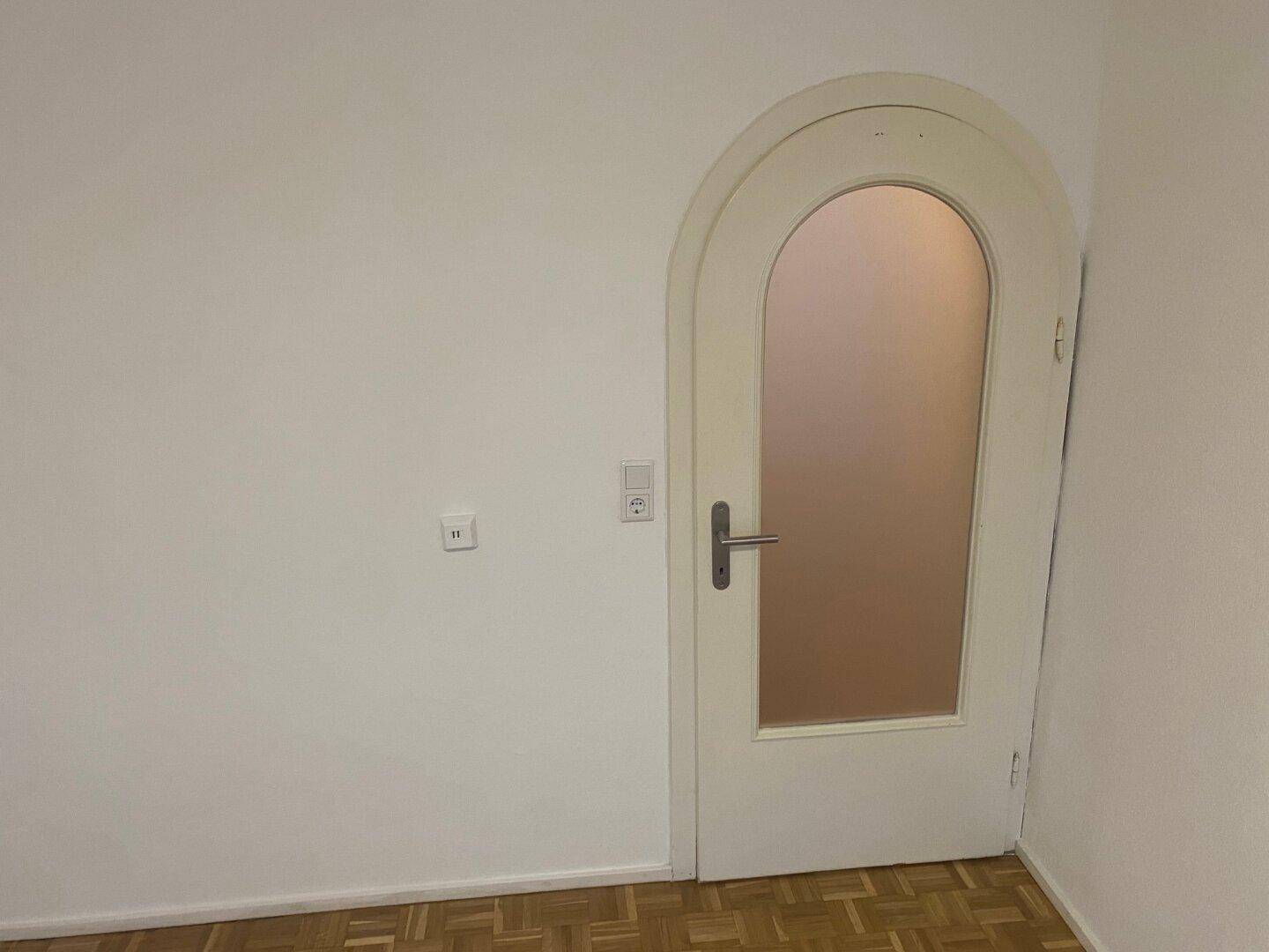 Weiße Wand mit einer bogenförmigen Tür aus Milchglas und Parkettboden, die zu einem anderen Raum führt.