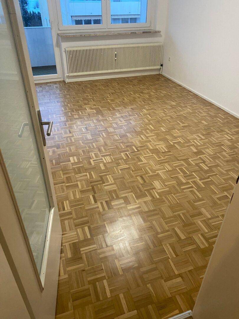 Helles, leeres Zimmer mit Parkettboden, großem Fenster und Heizkörper, bietet Zugang zu einem Balkon.