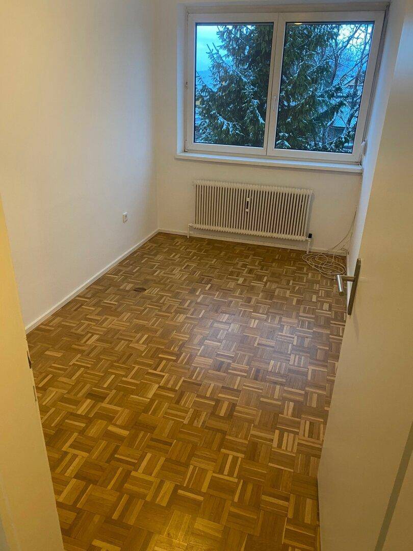 Leeres Schlafzimmer mit Parkettboden, weiß gestrichenen Wänden und einem Fenster mit Blick auf verschneite Bäume.