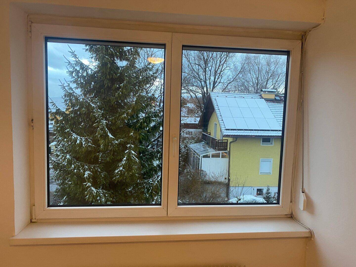 Fenster mit weitem Blick auf verschneite Bäume, ein Haus mit Solaranlage und die umliegende Berglandschaft.