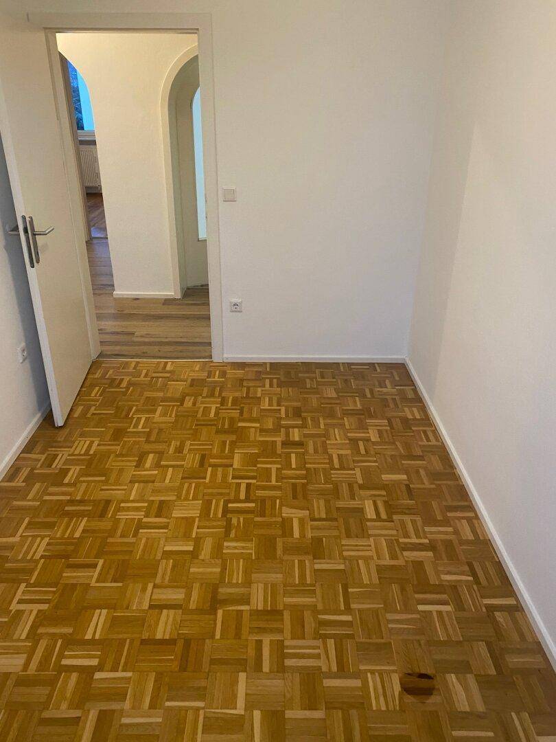 Helles, leeres Zimmer mit Parkettboden und weiß gestrichenen Wänden, das zu einem Flur führt.