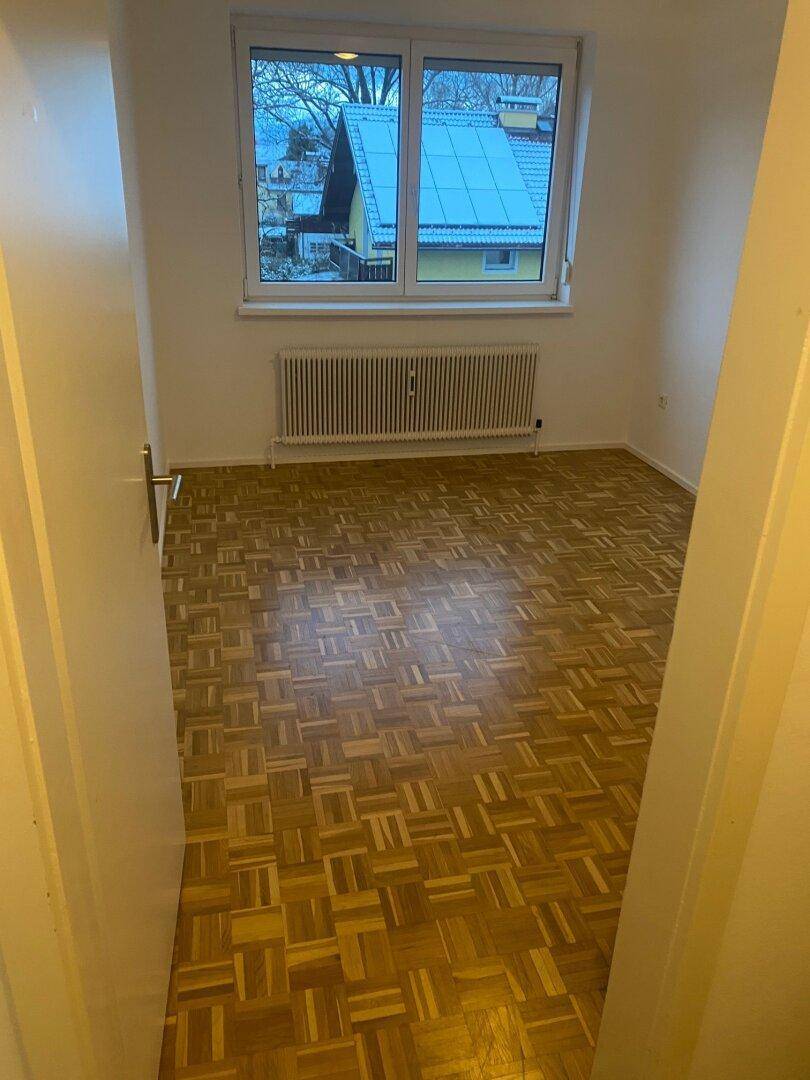 Leeres Schlafzimmer mit Parkettboden und Fenster, das einen Blick auf ein Haus mit Solaranlage bietet.